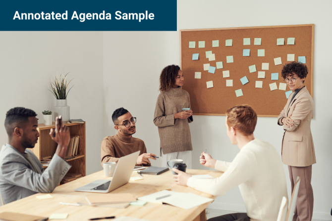 Photo of three people sitting at a long table and two people standing in front of a cork board with dozens of sticky notes attached to it